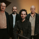 Julian Lage Quartet with John Medeski, Jorge Roeder, & Kenny Wollesen standing behind Julian Lage who is sitting and holding an electric guitar.