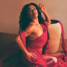 Lila Downs wears a flowing red outfit and poses near a large clay pot.