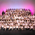A large group of girls in matching outfits posing together on stage with a pink backdrop.