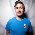 Mohanad Elshieky smiles at the camera in a bright blue t-shirt against a graffiti-covered wall.