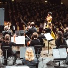 A full orchestra sits on a concert hall stage, applauding a soloist in a bright yellow dress who stands at center near a grand piano. Musicians in formal black attire hold violins, cellos, and other instruments, while a conductor stands to the side. Rows of audience members fill the background, creating a formal classical performance setting.  Is this conversation helpful so far?
