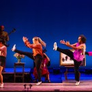 Four dancers mid-kick on blue-lit stage in colorful outfits, with a guitarist and cellist behind.