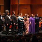 A group of six vocal performers on stage, dressed elegantly, with an orchestra in the background.