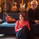 Rosanne Cash sits in front of John Leventhal on blue couch in guitar-filled studio