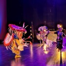 Three dancers in colorful feathered regalia performing on a purple-lit stage, photo