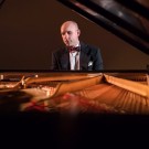 Christopher Taylor in suit and bow tie at grand piano, seen through open piano strings (photo)