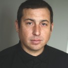 Headshot portrait of Tommy Orange with short dark hair, neutral expression, wearing dark shirt.