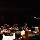 Conductor raising a baton to lead a full orchestra on a dimly lit concert stage.