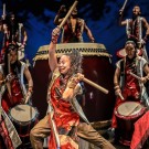 Energetic taiko drummers in red costumes performing with large drums on stage.