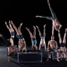 Acrobats forming handstands and midair flips above a box on a dark stage