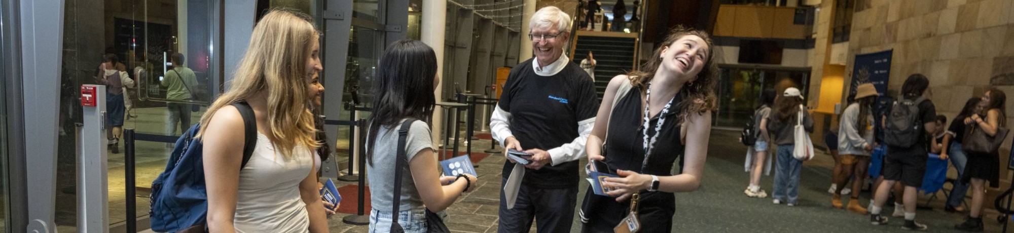 A small group of people chat and laugh inside a spacious, modern lobby with glass walls and high ceilings. Several young adults stand together holding phones or pamphlets while an older adult speaks with them, suggesting a welcoming or informational moment at a public event or campus venue.