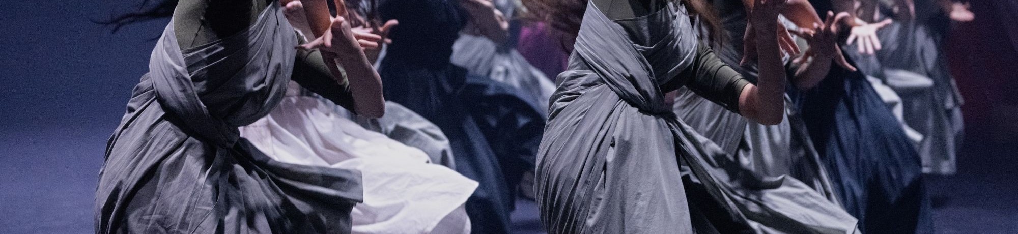 Dancers in flowing gray and white dresses in dynamic contemporary performance on dim stage