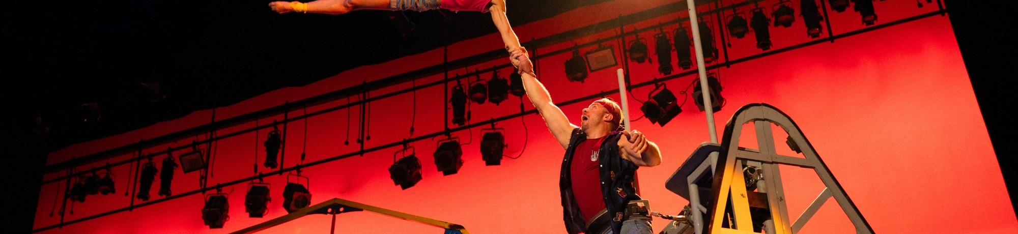 Man balances woman with one arm high above on red-lit circus stage.
