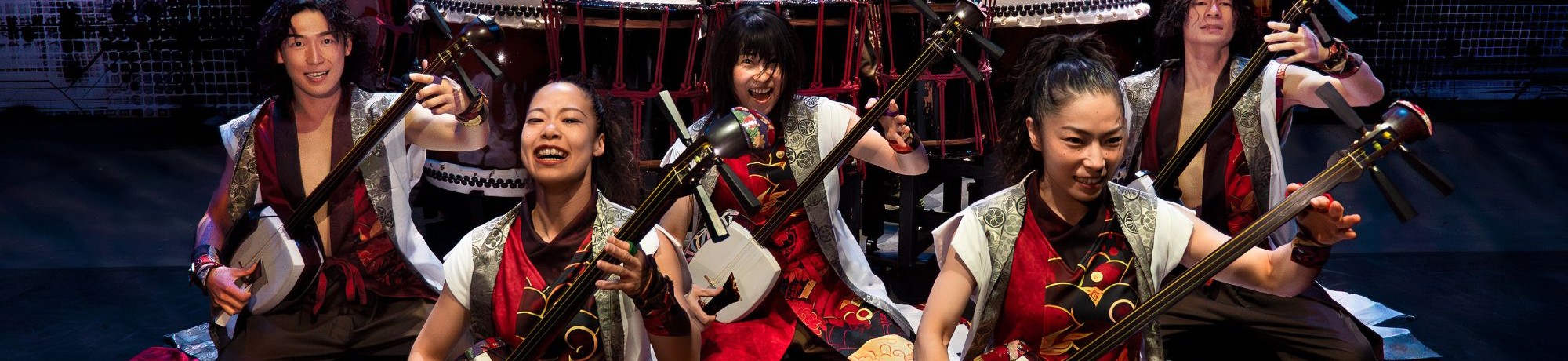 Nine Yamato performs in red-gold costumes playing taiko drums and stringed instruments.