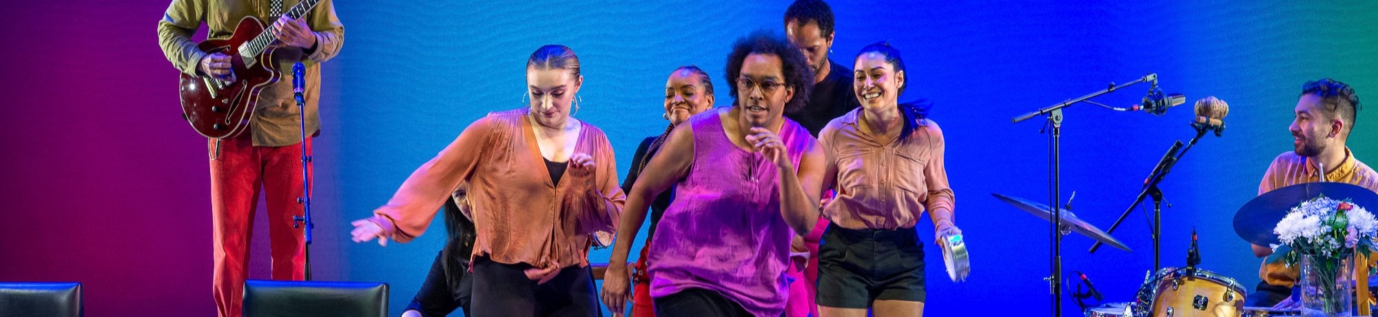 Dancers in bright shirts perform on a blue-green stage with musicians behind them