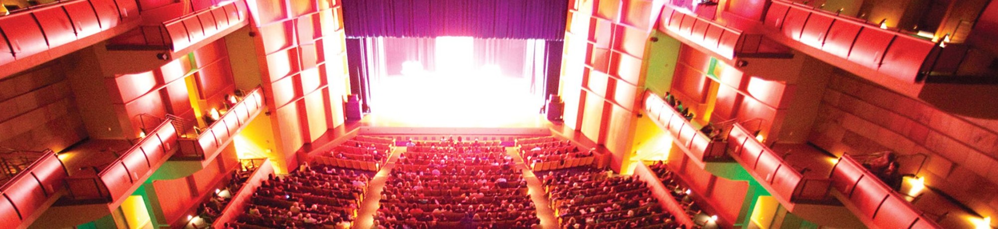 Vibrant theater interior with a large stage and audience, illuminated in colorful lights.