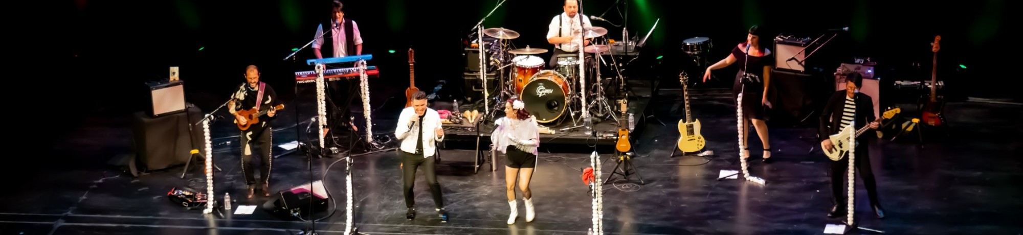 Las Cafeteras performs on a theater stage under green vertical lights. Multiple musicians are spread across the stage playing drums, keyboards, guitars, bass, and percussion, while two lead vocalists stand at center with microphones. Instruments, amps, and mic stands line the stage, creating the feel of a lively live concert performance.