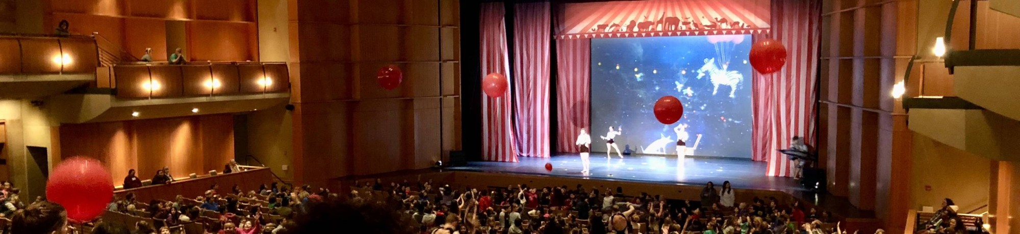 A crowded theater with people watching a performance on stage decorated with red balloons.