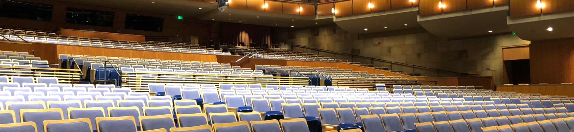 Interior view of Jackson Hall,with rows of empty blue and gray seats facing the stage. Multiple balcony levels with warm lighting and curved railings rise above the main floor, highlighting the spacious design of the performance venue before an event begins.