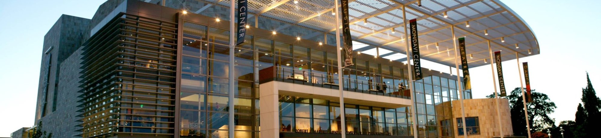 Modern building with a large glass facade and an expansive roof, illuminated at dusk.