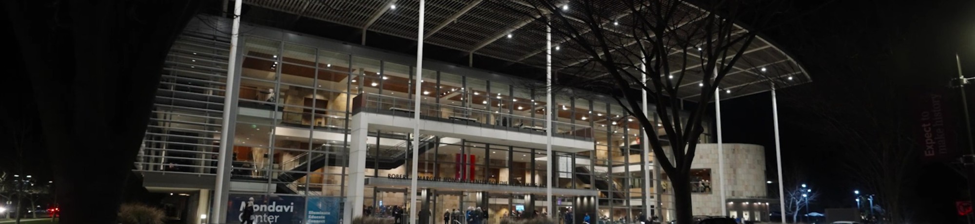 Exterior view of Mondavi Center at night, illuminated from within by warm lights. Glass walls reveal interior staircases and gathering spaces, with people visible near the entrance. Tall columns, a wide overhanging roof, and surrounding trees frame the Mondavi Center plaza after dark.