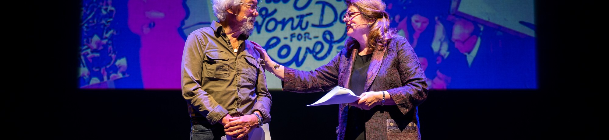 Two people on stage engage in conversation, with chairs and a colorful backdrop.